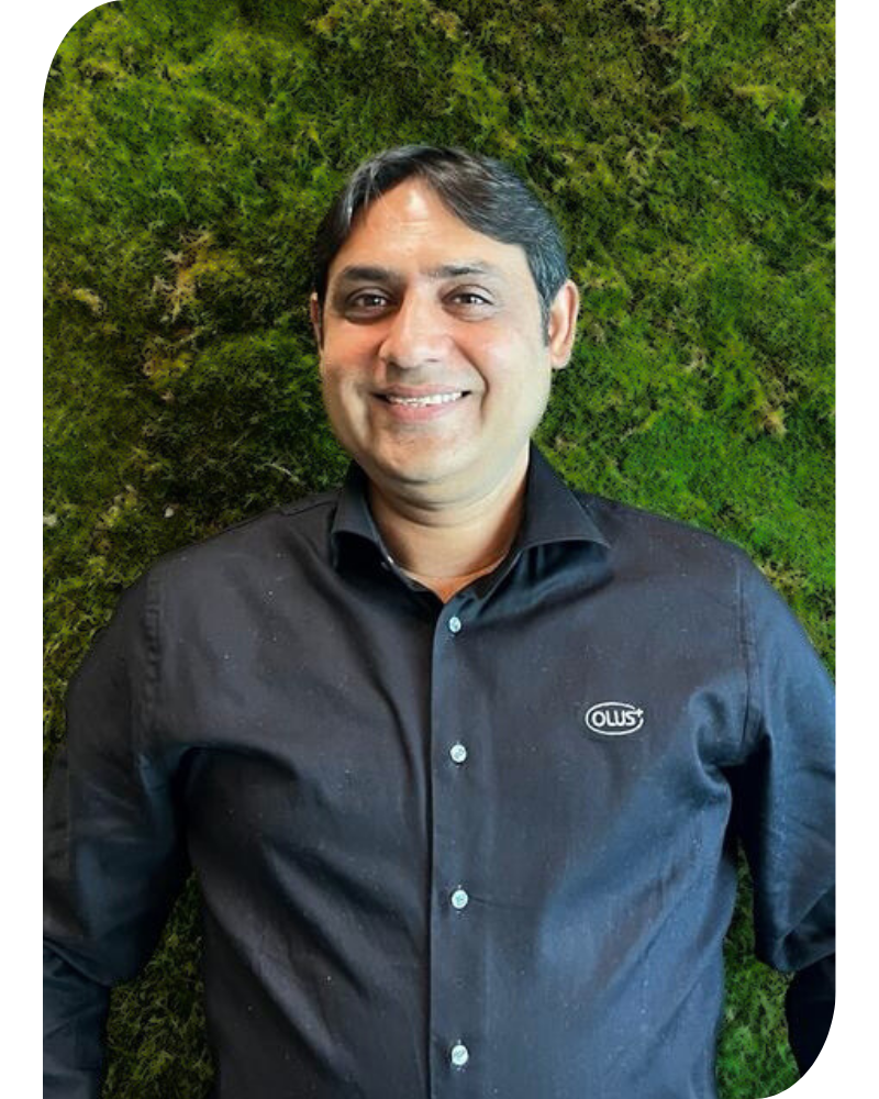 A photo of a smiling Pakistan male sales manager with dark hair, wearing a black blouse with the logo of Olus. Standing before a sustainable green moss wall. 